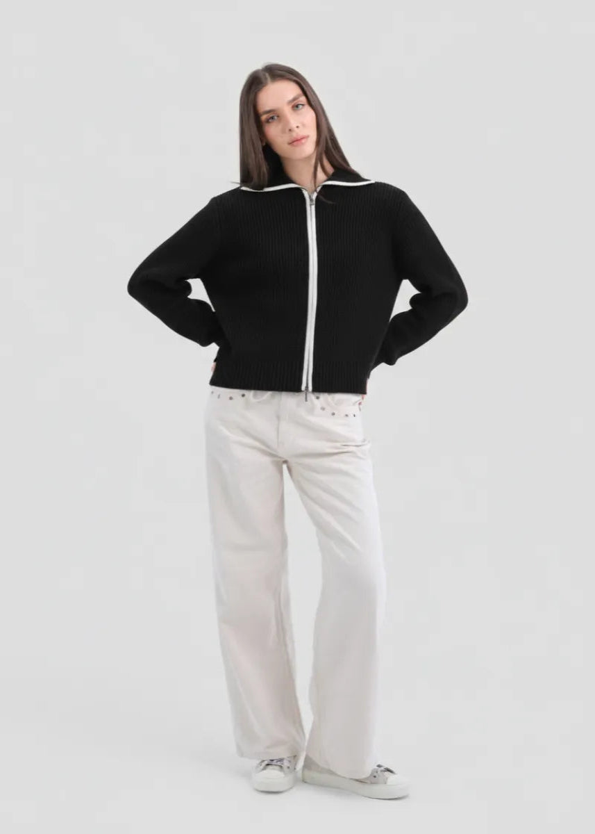 Woman wearing a black zip-up sweater and white pants on a light gray background