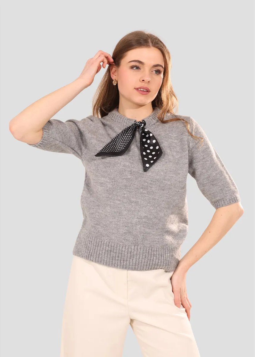 Woman wearing a gray sweater with a black polka dot scarf, standing against a light gray background