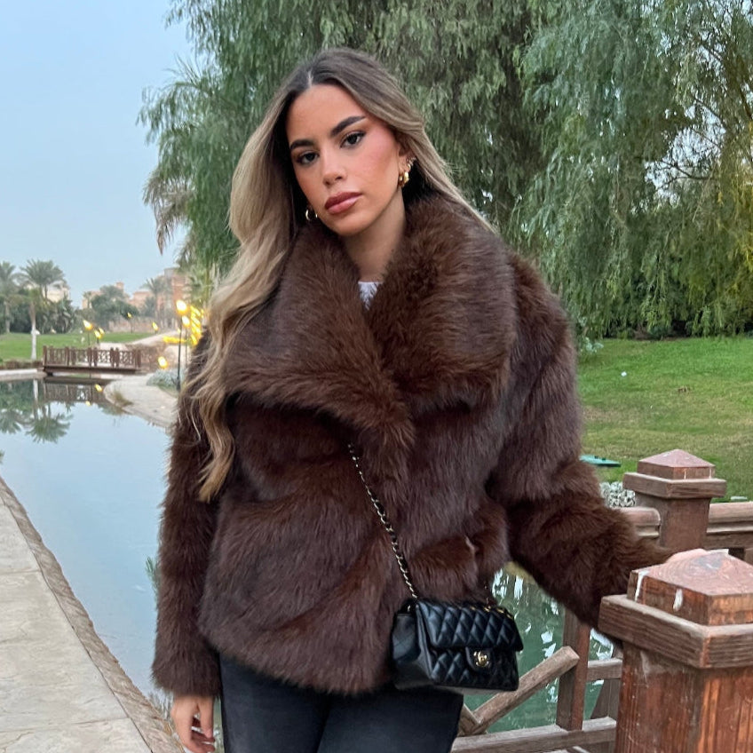 Woman in a brown fur coat standing by a poolside with trees in the background